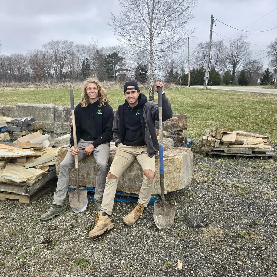 Two members of the Exterior Installations crew in company hoodies, sitting on a stone slab with shovels next to pallets of flagstone